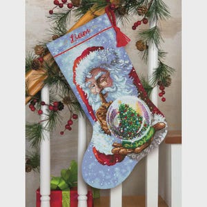 May include: A Christmas stocking featuring Santa Claus holding a snow globe with a Christmas tree inside. The stocking is blue with red accents and the name "Liam" embroidered in red. A red tassel hangs from the top.