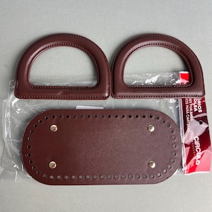 May include: A set of two dark brown semi-circular bag handles and one oval-shaped bag base with pre-punched holes and four gold-toned metal feet. The items are packaged in a clear plastic bag with a red and white label that reads CIRCULO.