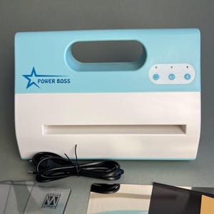 Puede incluir: Una plastificadora térmica Power Boss de color azul claro y blanco con asa superior, panel de control con tres botones circulares y cable de alimentación. Incluye fundas de plastificación transparentes y una hoja metálica protectora.