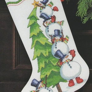 May include: A white Christmas stocking featuring a cross-stitched design of a green pine tree and four snowmen stacked vertically. The snowmen wear blue top hats, red mittens, and colourful scarves. The name MICHAEL is stitched in red at the top.