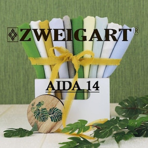 May include: A collection of Zweigart Aida 14 fabric pieces in various colors including green, yellow, white, and blue, arranged in a white container tied with a yellow ribbon. A round wooden disc with a green leaf print is placed in front.