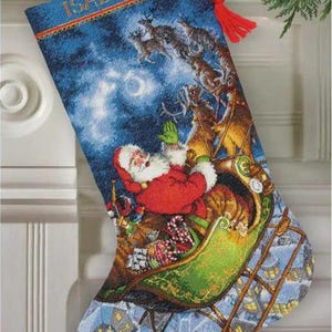 May include: A blue Christmas stocking featuring a cross-stitched scene of Santa Claus in a green sleigh pulled by reindeer against a starry night sky. The name ISABELLA is embroidered in orange at the top, and a red yarn tassel hangs from the side.
