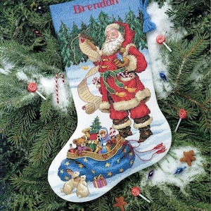 May include: A white Christmas stocking featuring a cross-stitched design of Santa Claus holding a long list, with a blue toy-filled sack at the base. The name Brendan is embroidered in red at the top, and a blue yarn loop is attached to the side.