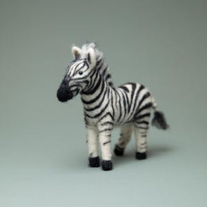 May include: A small, hand-felted zebra figurine with black and white stripes. The zebra is standing on a light gray background.