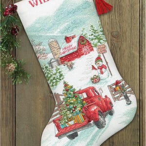 May include: A white fabric Christmas stocking featuring a cross-stitched winter scene with a red barn, a snowman, and a red pickup truck carrying a decorated Christmas tree. The name WILLIAM is embroidered in red at the top with a red hanging tassel.
