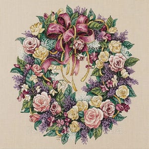 Op de afbeelding: Een kruissteek bloemenkrans met roze, gele en witte rozen, paarse seringen en groene bladeren. Een grote, donkerroze strik met gouden linten siert de bovenkant. De krans staat tegen een neutrale achtergrond.