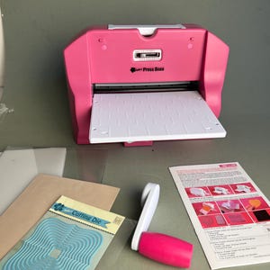 Puede incluir: Una máquina de troquelado y relieve Press Boss rosa y blanca con una bandeja de alimentación blanca, una manivela rosa y blanca desmontable, un paquete de troqueles de metal azul, placas de plástico transparente y una guía de instrucciones.