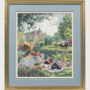 May include: Framed cross-stitch artwork depicting a serene picnic scene. The image shows a bridge, a river with a boat, and a group of people enjoying a picnic. The artwork is framed in a gold-colored frame with a blue mat.