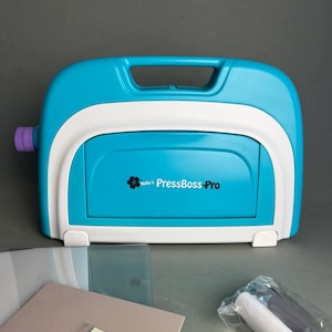 May include: A bright turquoise and white Nellie's PressBoss-Pro die cutting and embossing machine with a top handle. Included are several rectangular cutting plates in clear and light pink, along with a small grey tool in a plastic bag.