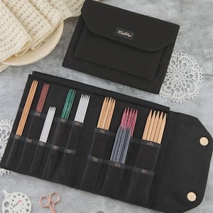 Könnte beinhalten: Eine schwarze Stricknadel-Tasche aus Stoff mit Stricknadeln aus Holz und Metall in verschiedenen Größen. Die Tasche ist geöffnet und zeigt die Nadeln in einzelnen Taschen. Eine kleinere schwarze Tasche und eine weiße Spitze sind ebenfalls sichtbar.