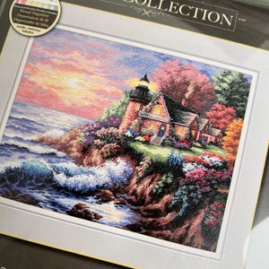 May include: A cross-stitch kit from The Gold Collection featuring a coastal scene with a lighthouse, house, and ocean waves. The image has a colorful sunset, with the text "Man of the Sea" at the bottom.