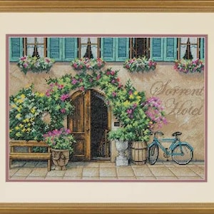 May include: Cross-stitch artwork depicting a building facade with the text "Sorrent Hotel". The scene includes a wooden door, a blue bicycle, and flower boxes. The artwork is framed in a gold-coloured frame.