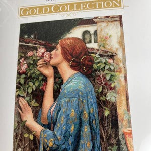 Op de afbeelding: Een Dimensions Gold Collection kruissteekpakket met een vrouw die aan een roos ruikt. De vrouw draagt een blauwe badjas met patroon. De kit bevat de tekst "DIMENSIONS" en "GOLD COLLECTION". De achtergrond toont een gebouw en bladeren.