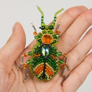 May include: A beaded insect brooch in shades of green, orange, and gold. The body is primarily green with orange accents, and the head features a large orange gem. The brooch is embellished with various beads and a central, teardrop-shaped crystal.