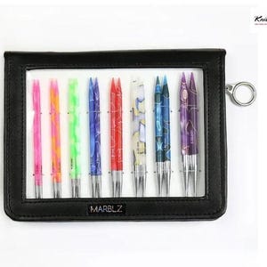 May include: A black case with a clear window containing a set of knitting needles in vibrant colours. The needles are in shades of pink, green, blue, and red. The case has a silver ring and the word "MARBLZ" is printed on the case.