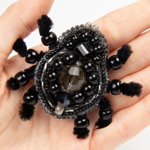 Puede incluir: Broche de araña negra con cuentas, con una gema central facetada y patas con cuentas. El cuerpo de la araña está adornado con cuentas negras y una variedad de otras gemas. El broche se sostiene en una mano.