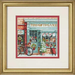 May include: A framed cross-stitch artwork depicting a storefront with the text TOYS-GIFTS-GAMES. The scene features a bicycle with a basket, a decorated Christmas tree, and a red sled. The storefront is painted in light blue with red accents.