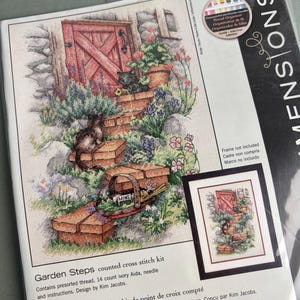 May include: A Dimensions Garden Steps counted cross stitch kit featuring a printed illustration of a red wooden door, stone steps, two cats, and a basket of mint. The package includes presorted thread, 14 count ivory Aida fabric, a needle, and instructions.