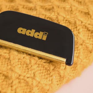 May include: A black fabric shaver with a gold-colored metal edge and the word addi printed in gold lettering on the front. The tool features a small heart icon integrated into the letter i of the brand name, designed for removing lint from textiles.