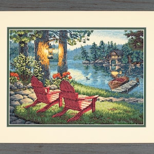 Könnte beinhalten: Kreuzstich-Kunstwerk einer Seeufer-Szene mit zwei roten Adirondack-Stühlen auf einem grasbewachsenen Ufer. Ein Holzsteg, ein Boot und eine Hütte sind über dem Wasser zu sehen. Das Kunstwerk ist in einem grauen Holzrahmen gerahmt.