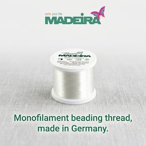 May include: A spool of clear monofilament beading thread by Madeira, featuring a white top label with the text Monofil, Madeira, Made in Germany, and various care symbols. The brand logo with a butterfly graphic is visible above the spool.