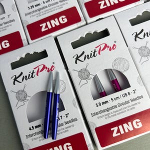 Könnte beinhalten: Mehrere Packungen KnitPro Zing austauschbare Rundstricknadeln in verschiedenen Größen. Jede weiße Schachtel zeigt das Markenlogo, einen kreisförmigen Ausschnitt mit zwei farbigen Metallnadelspitzen und Textangaben zur Größe in mm und US-Einheiten.