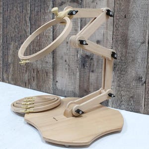 May include: A light-colored wooden embroidery hoop stand featuring an adjustable multi-jointed arm and a circular hoop attached at the top. A stack of four additional circular wooden embroidery hoops with gold-toned metal tightening hardware sits on the base.