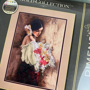 以下が含まれることがあります： Dimensions The Gold Collectionのクロスステッチキット。オフショルダーの白いブラウスとダークな花柄のスカートを身にまとい、色鮮やかな大きな花束を抱えたダークヘアの人物の肖像画が、茶色の背景に描かれています。