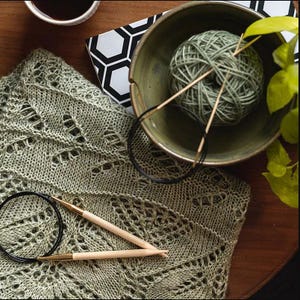 May include: A green knitted shawl with a delicate lace pattern, two wooden knitting needles, and a bowl of green yarn.