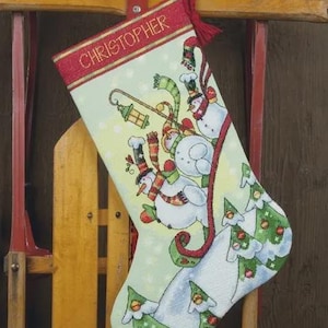 Puede incluir: Un calcetín navideño de color verde claro con el nombre CHRISTOPHER bordado en dorado en la parte superior. El calcetín muestra una escena festiva con tres muñecos de nieve, una linterna colgante y varios pinos con adornos rojos.