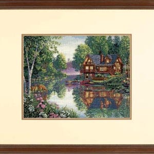 May include: A framed cross-stitch embroidery depicting a multi-storey log cabin situated on the edge of a calm lake surrounded by dense green trees. The scene includes a small red boat docked at the shore, pink flowers in the foreground, and a reflection of the cabin on the water surface, all enclosed within a dark brown wooden frame with a cream-coloured mat.