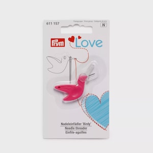 Puede incluir: Un enhebrador de agujas rosa con forma de pájaro sobre un cartón blanco. El empaque incluye el logo de Prym Love, el texto 'Nadeleinfädler Birdy', 'Needle threader', 'Einfile-aiguilles', un gráfico de línea roja discontinua y un corazón azul.