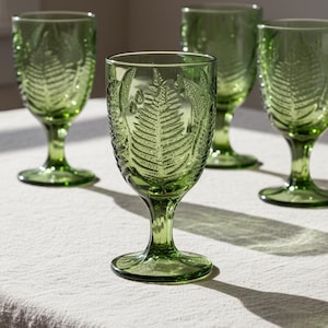 May include: Four green-tinted wine glasses with a fern leaf design. The glasses have a stem and a round base. The fern leaf design is etched into the glass. The glasses are arranged on a light-colored tablecloth.