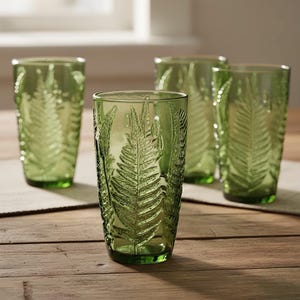 May include: Four green-tinted drinking glasses with a fern leaf design. The glasses are made of glass and are cylindrical in shape. The fern leaf pattern is embossed on the surface of the glasses. The glasses are sitting on a wooden surface.