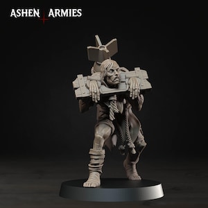 Pode incluir: Uma miniatura cinzenta da Ashen Armies representando uma pessoa presa num pelourinho de madeira. A figura tem cabelo comprido, veste roupas esfarrapadas com detalhes em corda e carrega um objeto grande tipo bomba nas costas sobre uma base.
