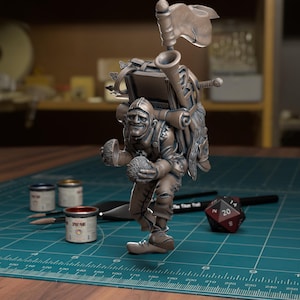 Puede incluir: Figura gris detallada de un personaje que lleva una mochila, una bandera y una bocina. La figura sostiene dos objetos redondos. Botes de pintura, pinceles y un dado están en primer plano. The Titan Troll está impreso en la superficie.