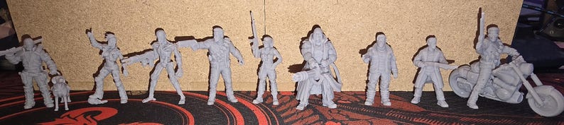 May include: A collection of eight unpainted gray miniature figures standing in a row. The set includes various human characters in tactical gear, some holding rifles or pistols, one accompanied by a small dog, and one figure mounted on a motorcycle.