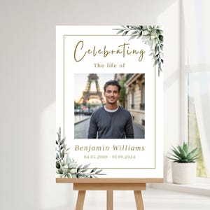 Puede incluir: Un letrero conmemorativo sobre un caballete con las palabras "Celebrating The life of" encima de una foto de un hombre frente a la Torre Eiffel. El letrero incluye el nombre Benjamin Williams y las fechas 04.05.2000 - 01.09.2024, con detalles florales.