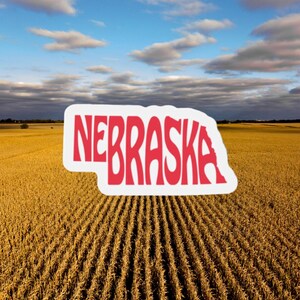 May include: A sticker in the shape of Nebraska with the word "NEBRASKA" in red letters. The sticker is set against a backdrop of a golden field under a cloudy blue sky.
