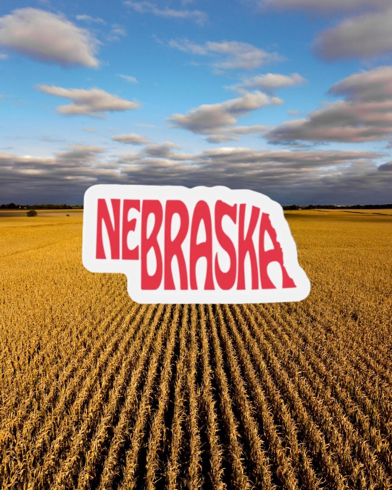 May include: A sticker in the shape of Nebraska with the word "NEBRASKA" in red letters. The sticker is set against a backdrop of a golden field under a cloudy blue sky.