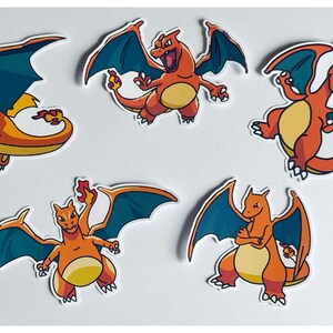 May include: Five orange and yellow Charizard stickers with blue wings are displayed. The stickers depict the Pokémon character in different poses, with detailed outlines and a white border. Ideal for personalising items like laptops, water bottles, or notebooks.