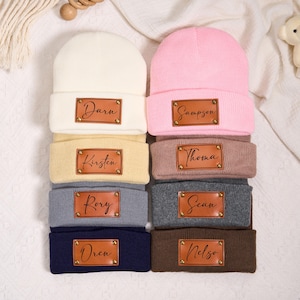 Può includere: Una pila di otto berretti lavorati a maglia in vari colori, tra cui crema, rosa, giallo, grigio, blu navy e marrone. Ogni berretto ha una toppa rettangolare in pelle con un nome. I nomi sono Darn, Sampson, Kirsten, Thoma, Rory, Sean, Oren e Nelso.