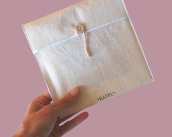 Può includere: Un pacchetto quadrato beige in stile busta con chiusura a bottone e spago rosa. Il pacchetto è tenuto in mano su uno sfondo rosa. La parola "RAPPU" è stampata nell'angolo in basso a destra.