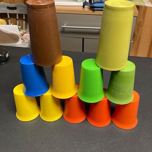 Puede incluir: Una torre de vasos de plástico de colores brillantes en tonos amarillo, azul, verde, naranja y marrón. Los vasos están apilados en forma de pirámide, destacando su tamaño y forma. Estos vasos probablemente son para juegos infantiles.