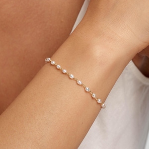 Puede incluir: Una delicada pulsera de cadena dorada que presenta una serie de pequeñas perlas blancas espaciadas uniformemente. La pulsera se lleva en la muñeca, mostrando los finos eslabones metálicos que conectan cada perla en un diseño simple y refinado.
