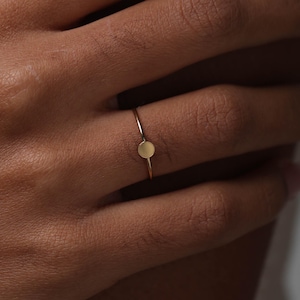 Può includere: Un delicato anello d'oro con un piccolo disco circolare come elemento centrale. La sottile fascia dell'anello e la superficie liscia del disco creano un design minimalista ed elegante. L'anello è mostrato su una mano, evidenziando la sua sottile bellezza.