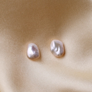 Pu&ograve; includere: Due orecchini di perle irregolari, bianco sporco. Le perle hanno una lucentezza lucida e una sfumatura leggermente rosata. Sono presentati su uno sfondo di tessuto beige satinato, creando un'estetica morbida ed elegante.