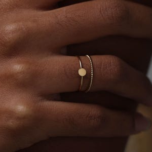 Puede incluir: Dos anillos dorados en un dedo. Un anillo presenta un pequeño círculo plano, mientras que el otro tiene un diseño de cuentas. Los anillos son delicados y minimalistas, con un acabado pulido. Los anillos se colocan sobre un fondo cálido y neutro.