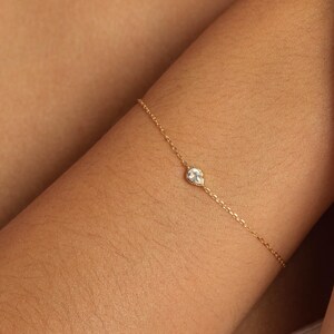 Op de afbeelding: Een delicate gouden kettingarmband met een enkele, kleine, peervormige iriserende edelsteen gezet in een gouden zetting. De armband is samengesteld uit fijne, gepolijste gouden schakels die een doorlopende, slanke band om de pols vormen.