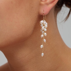 Peut inclure: Une boucle d'oreille pendante dorée composée d'une grappe de petites perles d'eau douce blanches de forme irrégulière, suspendues à une fine chaîne dorée. Les perles sont disposées en cascade, formant un design long, fin et texturé.